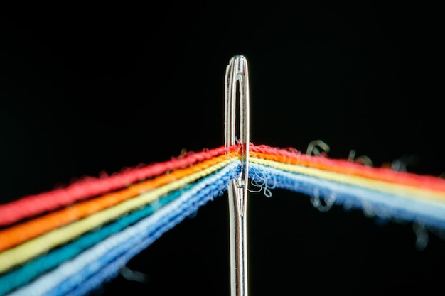 Multicolored threads for sewing in the form of a rainbow pass through an antique needle on a black background