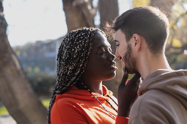 Multicultural couple of sweethearts in engaging moment with an African woman touching tenderly her boyfriend chin under the trees in the park