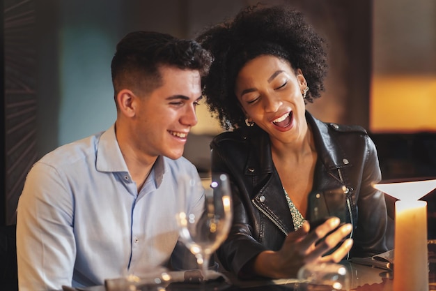Многокультурная пара молодых людей, использующих смартфон и социальную сеть, сидя за столом в ресторане.