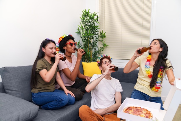 Multicultural friends eating pizza and toasting with beers on sofa at a relaxed birthday celebration