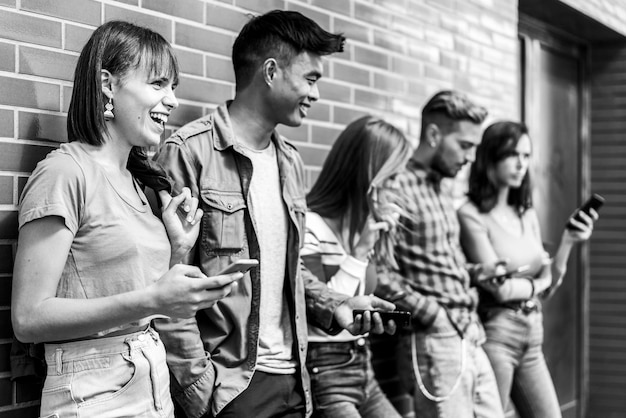 Photo multicultural friends laughing using smartphone at wall on university college campus young people addicted by mobile smartphones technology concept with always connected milenials vintage edit