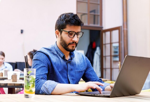 Multicultural smart student in jeans shirt browsing something at the laptop while sitting at the cafe