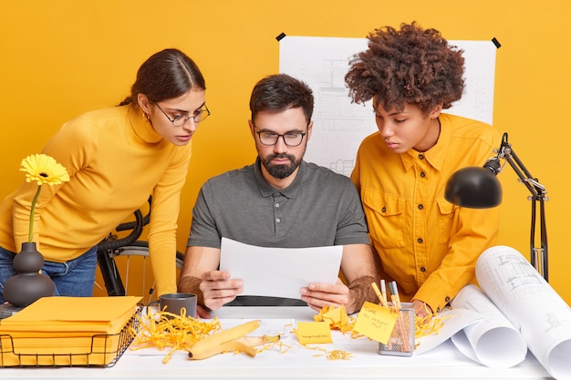 Multicultural team group collaborate together at workplace have brainstroming meeting in office focused attentively at paper try to decide something. Man architect with two female assistants