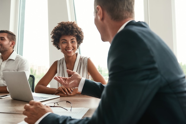 Multicultural team. Young and beautiful afro american woman discussing something with colleagues and smiling while sitting in the modern office. Brainstorming. Business meeting