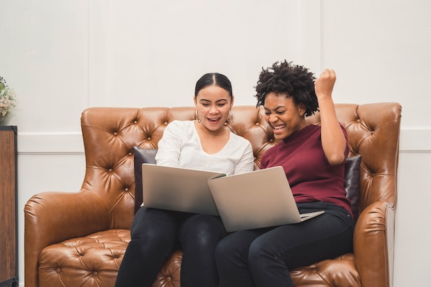 Multicultural women using a laptop on couch relaxing on the living room and laughing at something on the screen