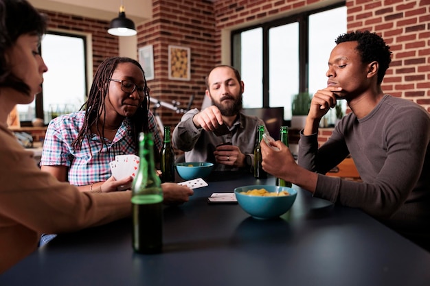 Multiculturele groep vrienden die aan tafel in de woonkamer zitten terwijl ze samen een gissend kaartspel spelen. multi-etnische jonge volwassen mensen die thuis genieten van leuke vrijetijdsbesteding.