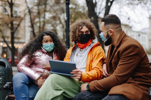 Multiculturele jongeren met medische maskers die samen vrije tijd op straat doorbrengen en een digitale tablet gebruiken. Gelukkige mannen en vrouwen die op houten bank met modern apparaat zitten.