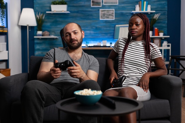 自宅のソファに座ってコンソールでビデオゲームをプレイする多民族の大人のカップル。テレビでコンソールゲームをしたり、お椀でおやつを食べたりして、自由な時間を楽しんでいるリラックスした人々。