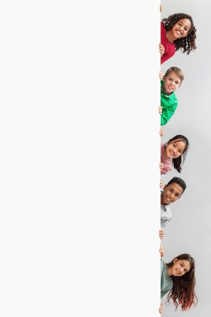 Multiethnic children hiding behind white board smiling over gray background