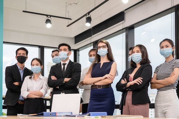 Multietnico fiducioso business team in piedi con il braccio incrociato e indossando la mascherina medica in ufficio moderno