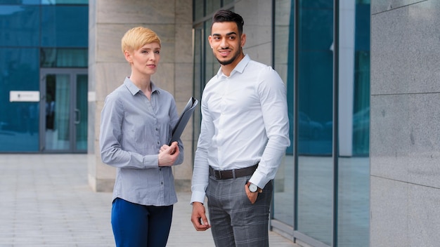 Multiethnic couple arabic indian business man and caucasian woman holding folder two colleagues partners standing outdoors taking break in work looking away waiting meeting