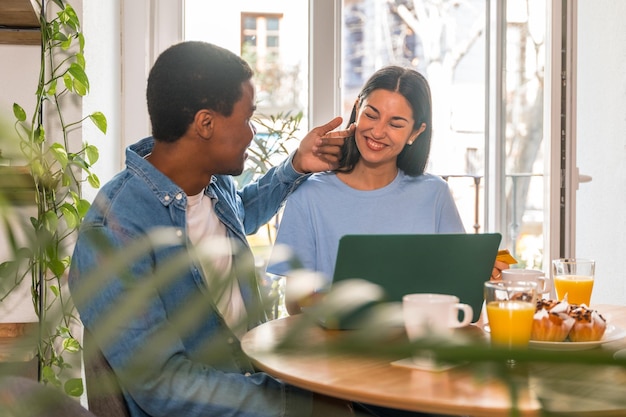 Multiethnic couple in love making online purchase with the computer while having breakfast by the window