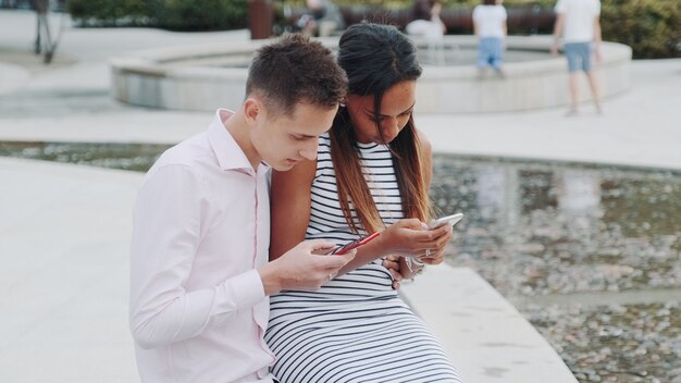 Multiethnic couple sitting together outdoors and using smartphones