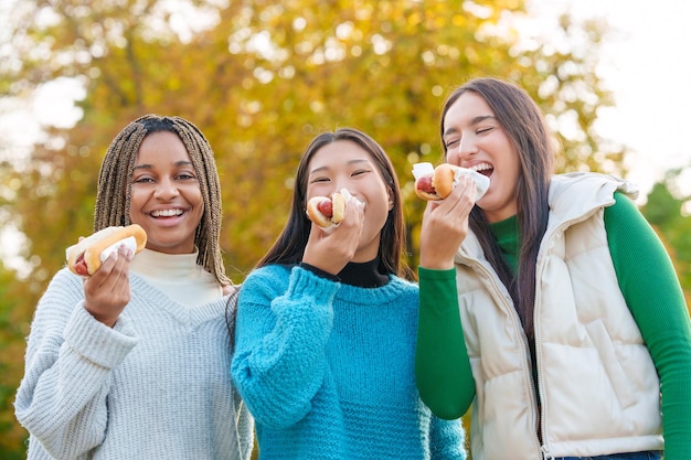 Photo multiethnic friends joking while eating hot dog in a park