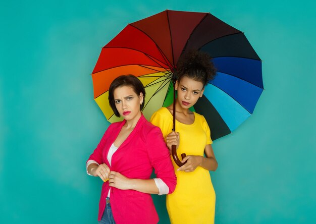 Multiethnic friendship concept. Mulatto and caucasian angry girls in colorful casual clothes posing under rainbow colored umbrella at blue studio background with copy space