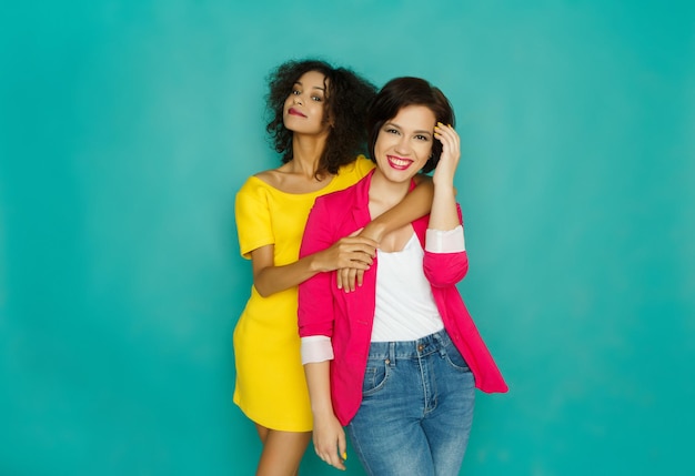 Multiethnic friendship and support concept. African-american and caucasian girls in colorful casual clothes hugging and having fun at blue studio background with copy space looking at camera