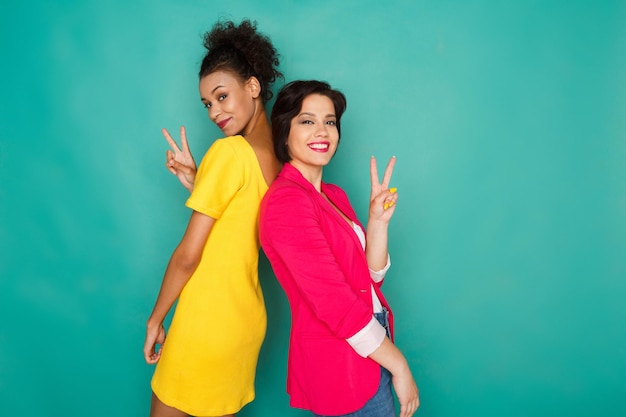 Multiethnic friendship and support concept. Happy mulatto and caucasian girls in colorful casual clothes, standing back to back showing victory sign at blue studio background with copy space