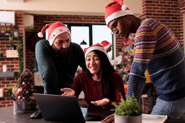 Multiethnic group of colleagues using laptop on christmas, working on project in office decorated with festive ornaments and xmas tree. People with santa hat doing teamwork during holiday.