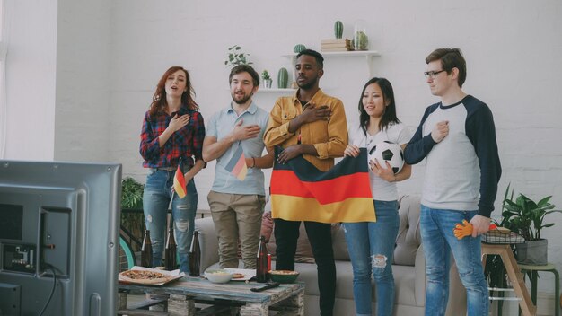 Multiethnic group of friends listening and singing german\
national anthem before watching sports championship on tv together\
at home