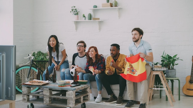 Gruppo multietnico di amici appassionati di sport con bandiere spagnole che guardano il campionato di calcio in tv insieme a casa e rallegrano la squadra del cuore