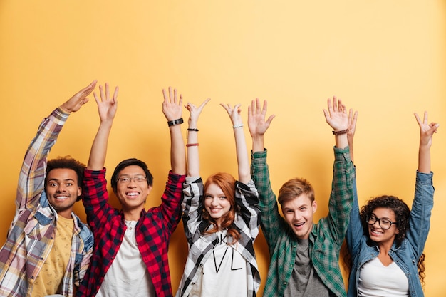 Gruppo multietnico di giovani felici in piedi e che celebrano il successo con le mani alzate su sfondo giallo