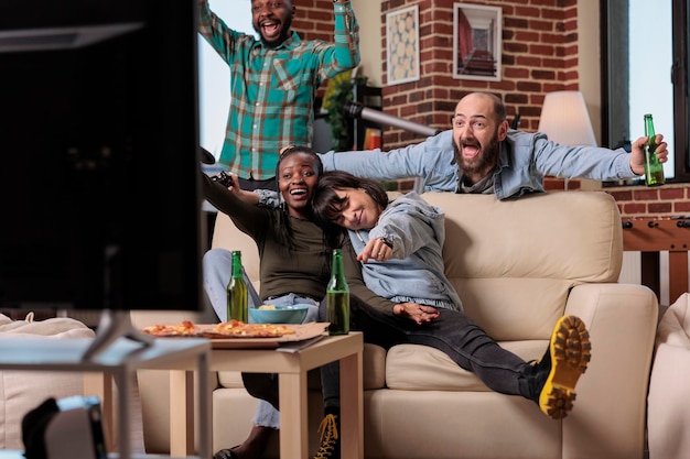 Gruppo multietnico di persone che celebrano i videogiochi vincono insieme, giocando a gare di festa con l'alcol. le persone si sentono felici per la vittoria e la vittoria, bevendo birra a casa.