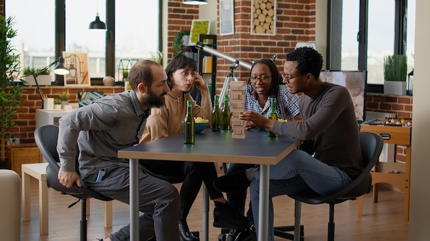 Multiethnic group of people having fun with tower pieces,\
playing board games with square building and wooden blocks on\
table. friends laughing and enjoying leisure pasttime\
activity.