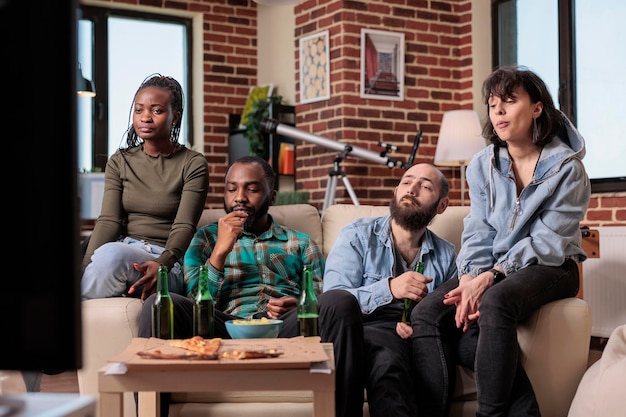 Multiethnic group of people watching movie on television, eating popcorn and drinking bottles of beer at fun gathering. People looking at film on tv channel program, enjoying leisure activity.