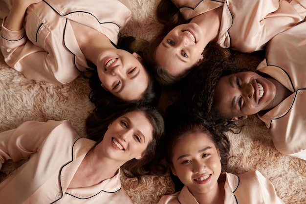 Многонациональная группа улыбающихся молодых женщин в шелковых пижамах, лежащих на полу