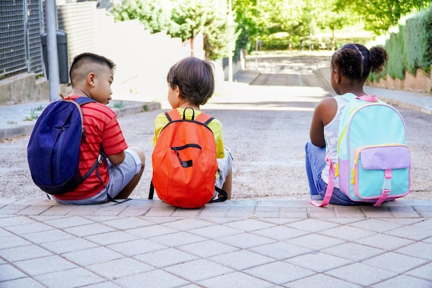 学校の入り口の通りに座ってりんごを食べているバックパックを持つ多民族の子供たち。