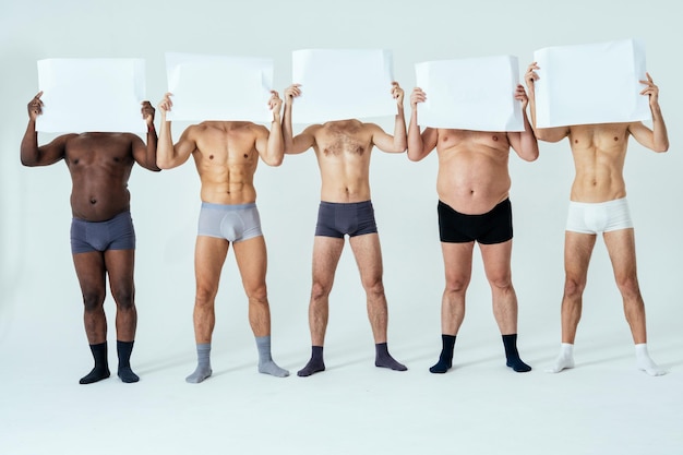 Photo multiethnic men posing for a male edition body positive beauty set showing messages on banners. shirtless guys with different age, and body wearing boxers underwear