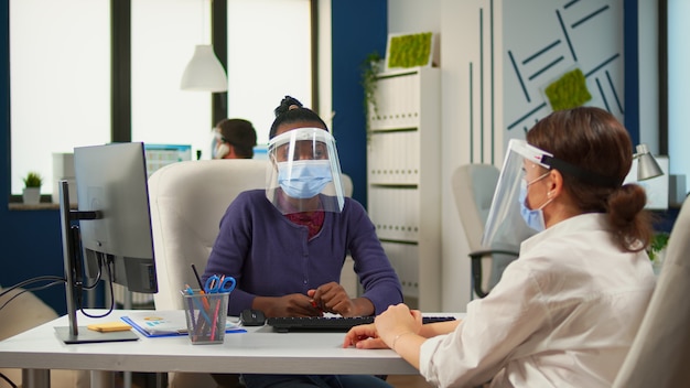 Multiethnic partners working in office with new normal sitting at desk in front on computer discussing strategy wearing protections masks. Group of diverse businesspeople respecting social distance.