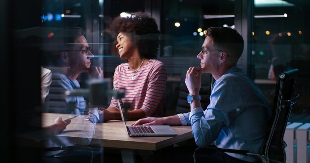 Мультиэтническая стартап-команда на встрече в современном ночном офисе проводит мозговой штурм, работает на ноутбуке