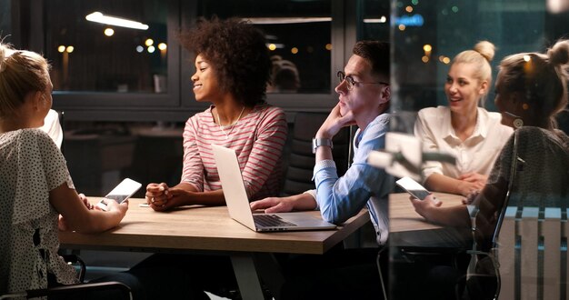 Мультиэтническая стартап-команда на встрече в современном ночном офисе проводит мозговой штурм, работает на ноутбуке