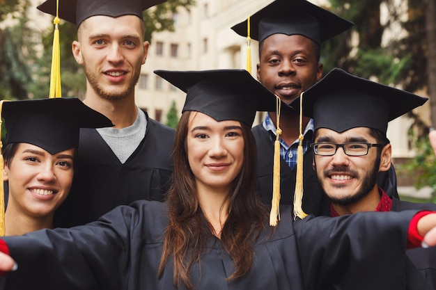 Multiethnic students taking group selfie on smartphone to celebrate their graduation. Education, qualification and gown concept.