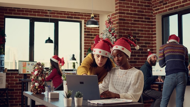 Photo multiethnic team of women planning research on laptop, working on startup project in office with christmas tree and lights. festive colleagues doing teamwork in space with xmas holiday decor.