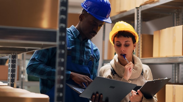 Multiethnic team of workers analyzing list on laptop and tablet, working with industrial cardboard boxes in storage room. People checking merchandise stock for delivery service. Handheld shot.