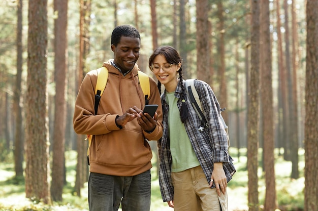 Multiethnic young couple using mobile phone while standing in the forest during hiking