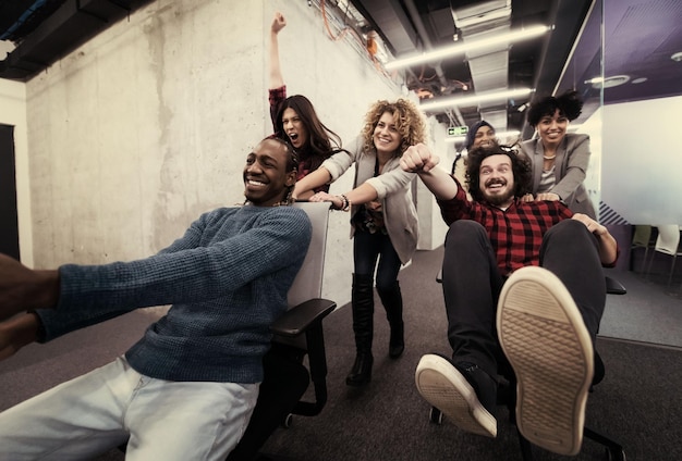 Startup multietnica team di sviluppatori di software che si divertono mentre corrono su sedie da ufficio, diversi dipendenti entusiasti che ridono godendosi attività divertenti durante la pausa di lavoro, lavoratori creativi e amichevoli p