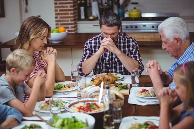 Multigeneratiefamilie die met grootouders bidden die bij eettafel zitten