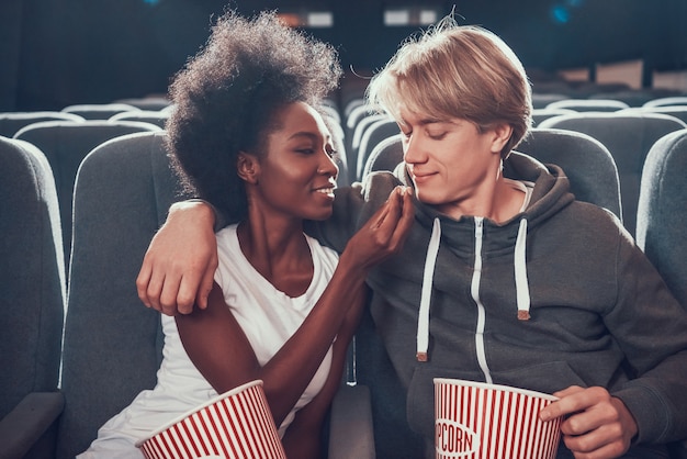 Multinational couple is feeding each other in cinema.