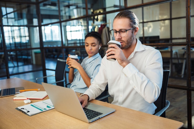 многонациональные серьезные коллеги работают с ноутбуками и пьют кофе, сидя за столом в офисе