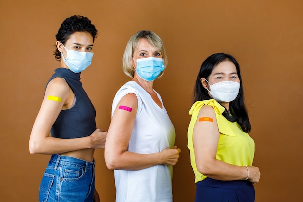 Multinationale etnische vrouwelijke patiënten dragen een gezichtsmasker dat in de hoogte staat, kijk naar de camera met kleurrijke pleisters samen na de injectie van de coronavirusvaccinatie voor de bruine muurachtergrond.