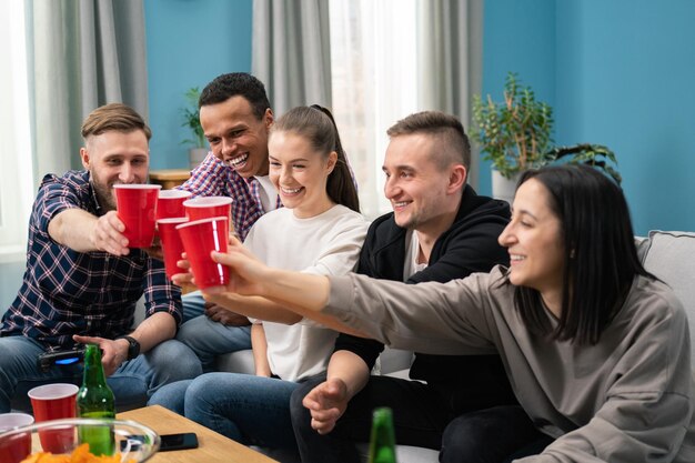Multinationale vrienden die een kleine thuisfeestbijeenkomst hebben, zittend op de bank met een rood kopje bier