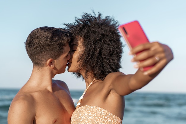Multiraciaal koppel dat selfie op het strand neemt en samen zoent tegen de zee