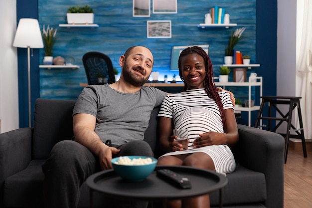 Multiraciaal paar dat van televisie geniet en thuis een baby verwacht terwijl u ontspant op de bank. Lieve mensen die zich gelukkig voelen bij het kijken naar een informele komedie samen op tv in de woonkamer terwijl ze wachten tot de baby wordt geboren