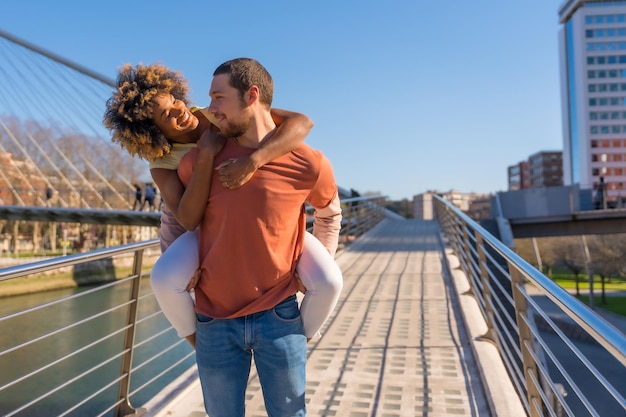 Multiracial couple on city street lifestyle smiling man with girlfriend on his back on catwalk