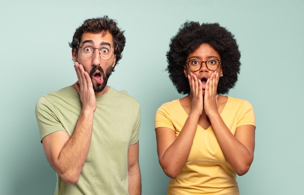 Multiracial couple of friends feeling shocked and scared, looking terrified with open mouth and hands on cheeks