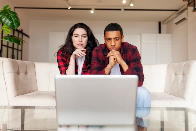 multiracial couple looking puzzled and serious at laptop on sofa