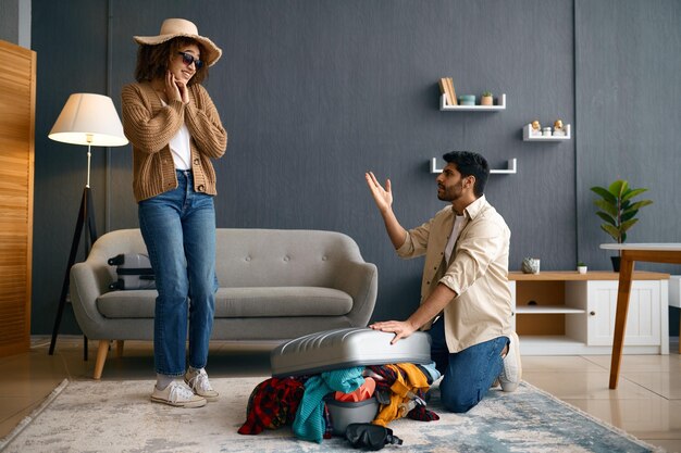 Multiracial couple packing clothes into travel suitcase. Husband refusing to take more clothes